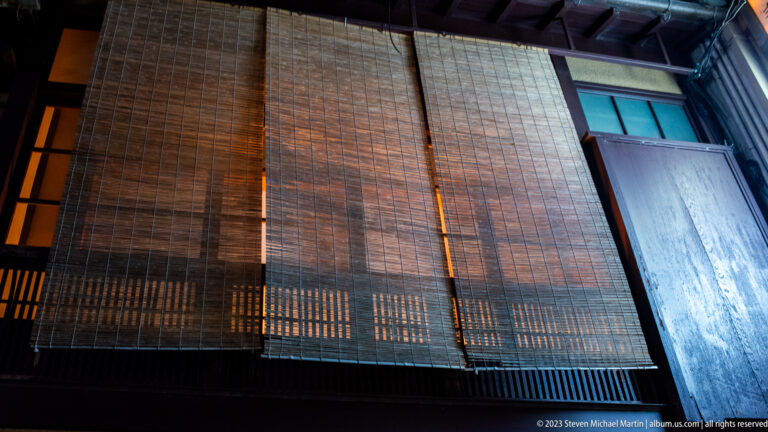 Traditional timber façade at night with large rolled bamboo blinds covering the upper level.