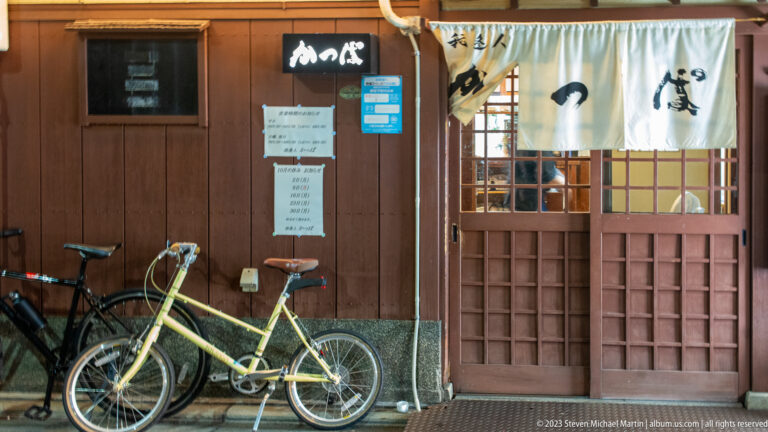 Small wooden restaurant with white noren, sliding doors, posted menus, and two bicycles outside.