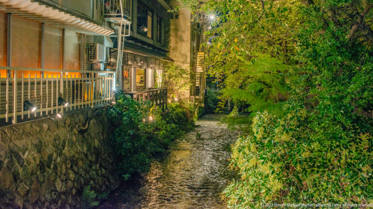 Narrow canal at night with stone banks, overhanging greenery, and lit restaurants beside the water.
