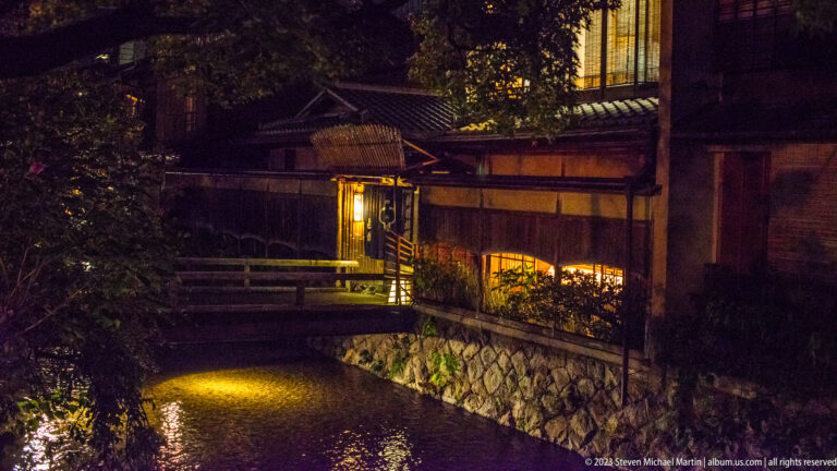 Dark Kyoto canal with warm-lit wooden buildings, a small footbridge, stone bank, and reflections.
