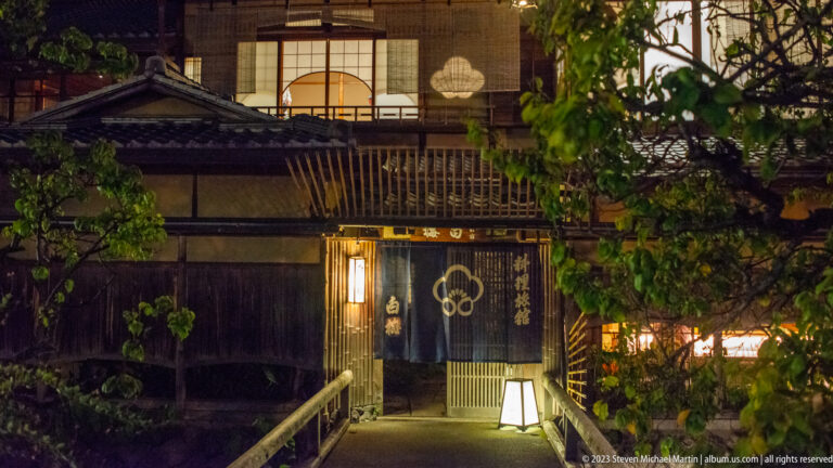 Narrow lit path leading to a traditional entrance with dark noren, latticework, and glowing upper windows.
