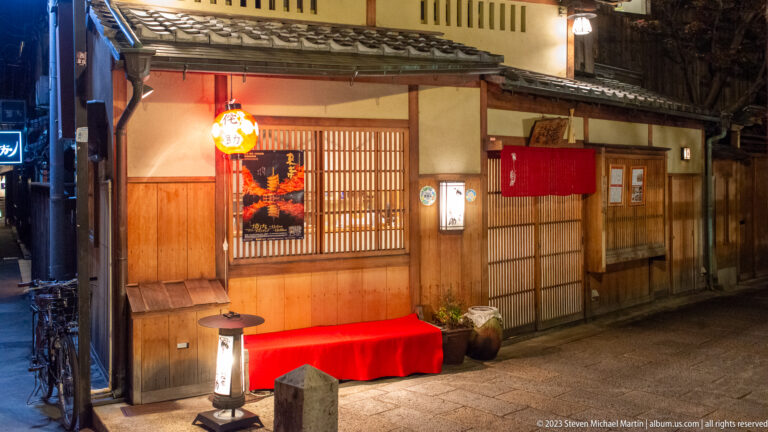 Traditional wooden restaurant with red bench, glowing lantern, lattice window, and red noren at night.