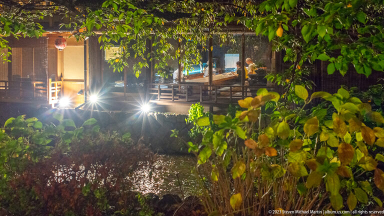 Dining terrace viewed through riverside foliage shown at night with the principal architectural, landscape, or street details visible in the composition.
