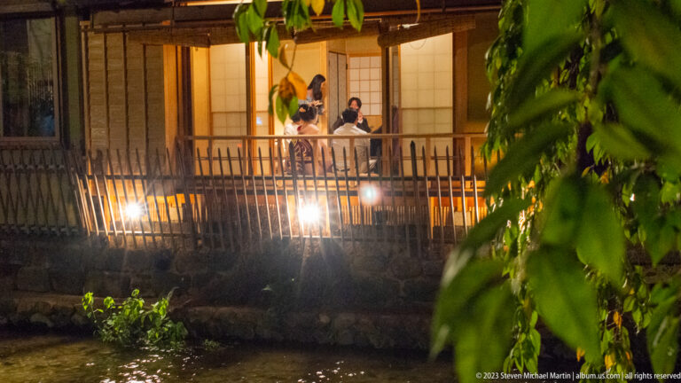 Diners seated on an upper veranda overlooking the canal shown at night with the principal architectural, landscape, or street details visible in the composition.