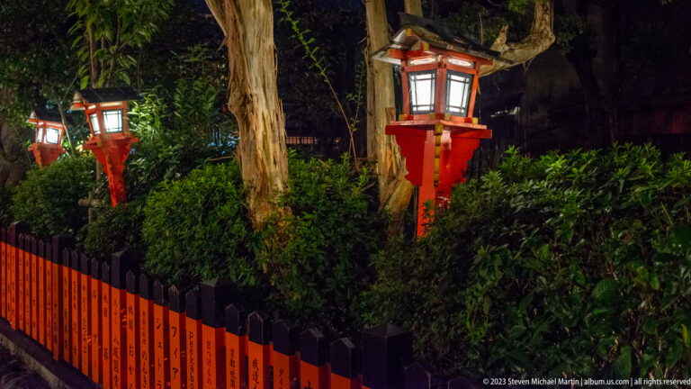 Vermilion shrine lanterns beside a sacred enclosure shown at night with the principal architectural, landscape, or street details visible in the composition.