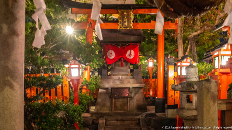 Small shrine beneath torii and lanterns shown at night with the principal architectural, landscape, or street details visible in the composition.