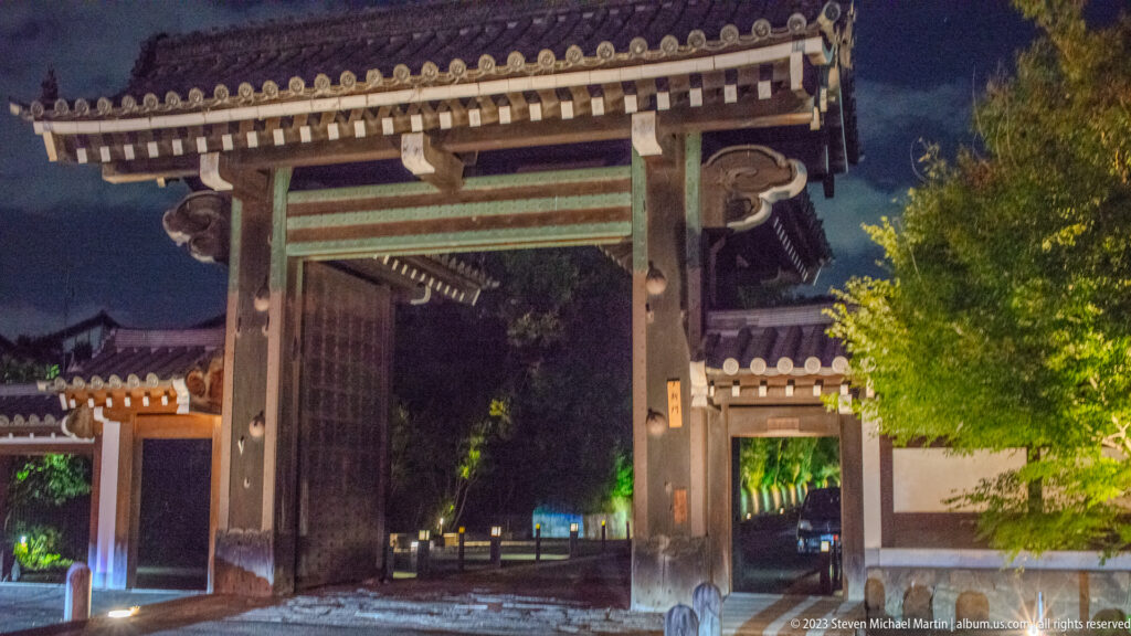 Large traditional Japanese gate at night with tiled roof, timber brackets, and an open passage.