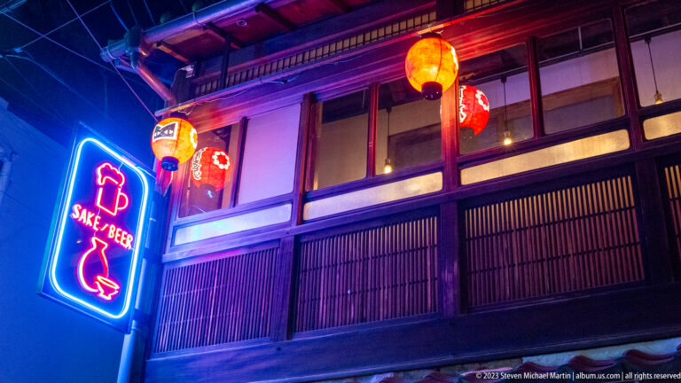 Neon sake-and-beer sign beside a traditional wooden Kyoto storefront with red paper lanterns.