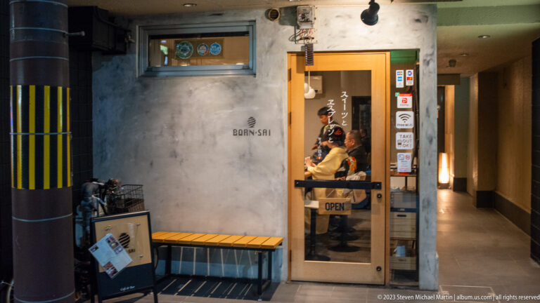 Narrow restaurant entrance with concrete walls, a wooden bench, menu board, and warm interior light.