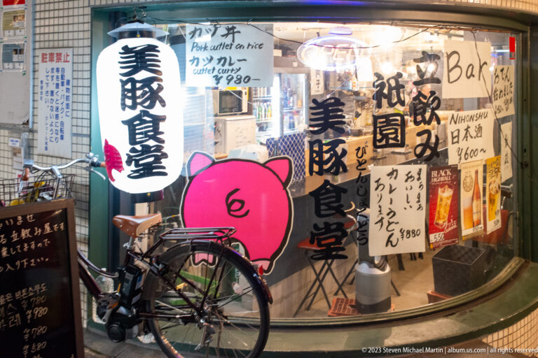 Japanese restaurant window covered with handwritten menus, bold lettering, a pink pig graphic, and a parked bicycle.