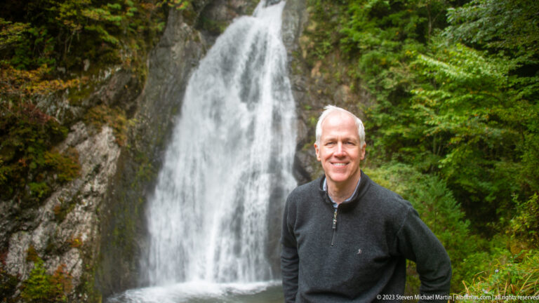Smiling man in a dark sweater posed beside Choshi Falls and green foliage.