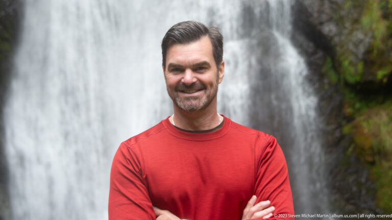 Dark-haired man in a red shirt with crossed arms before a blurred waterfall.