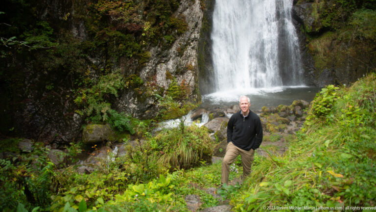 Small standing figure beside Choshi Falls in a steep, green rocky gorge.