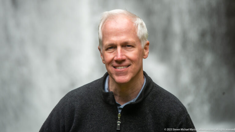 Close portrait of a smiling gray-haired man with a blurred waterfall behind him.