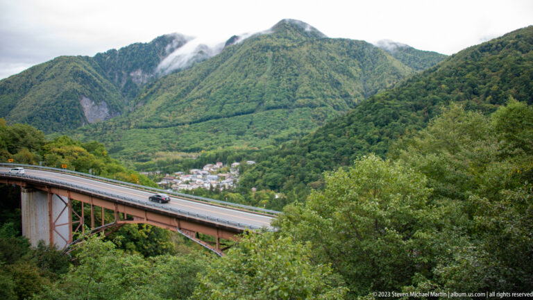 Steel road bridge crossing a forested mountain valley above a small settlement.