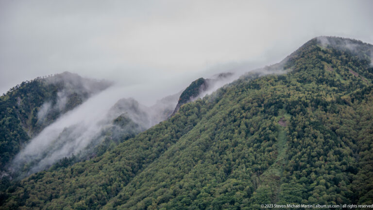 Low clouds wrapping around a steep, densely forested mountain ridge.