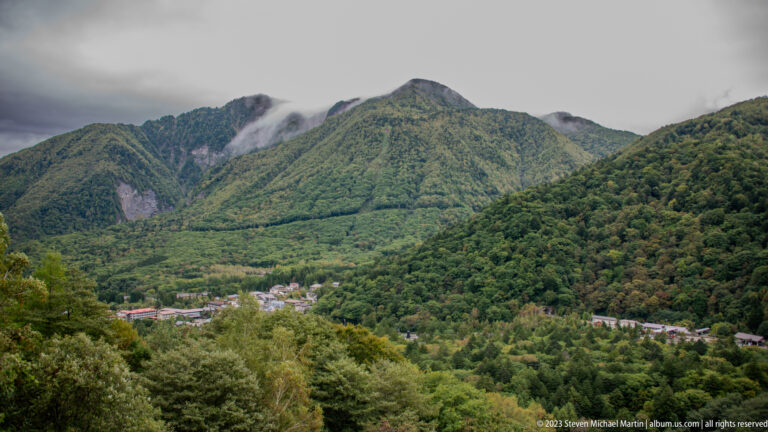 Village at the base of steep green mountains partly covered by low cloud.