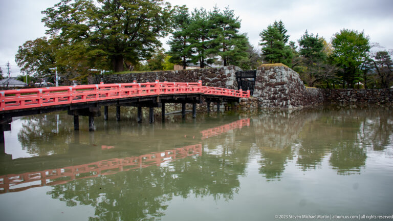 Red timber bridge reflected in Matsumoto Castle’s moat between stone abutments.