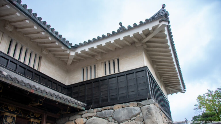 Low-angle detail of white plaster, black timber siding, tiled eaves, and stonework.