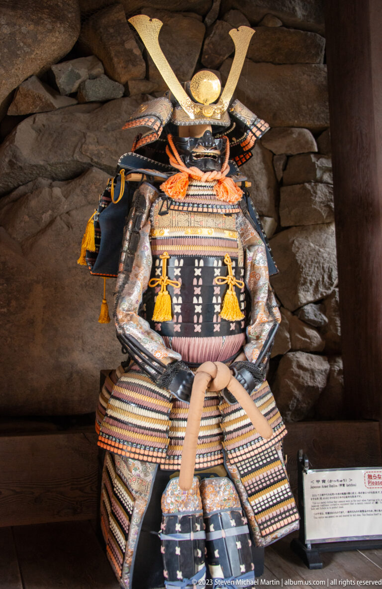 Japanese samurai armor with a crescent-crested helmet displayed against timber beams and a stone wall inside Matsumoto Castle.