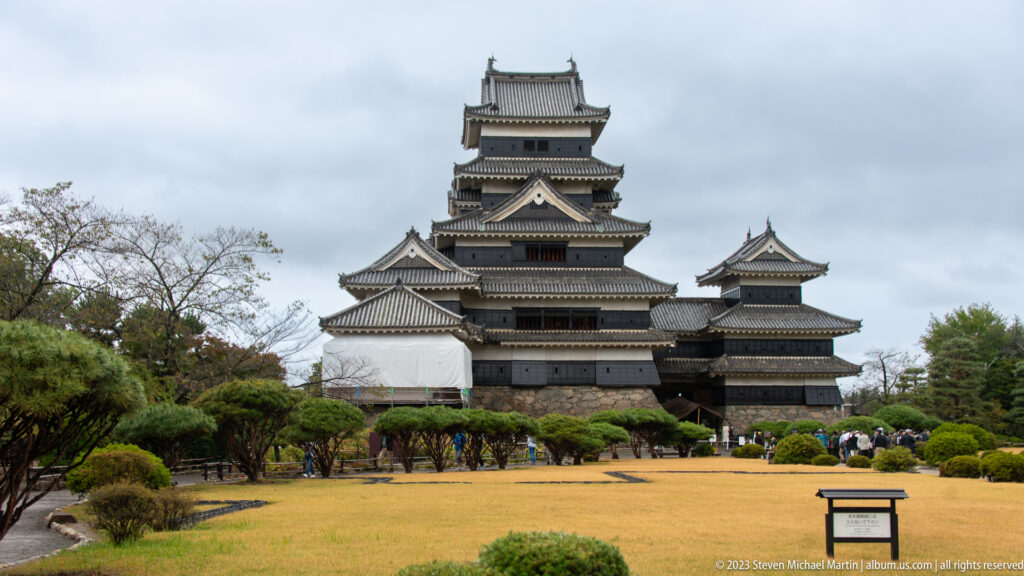 Matsumoto Castle’s main keep and adjoining towers seen across a lawn with clipped pines and visitors.