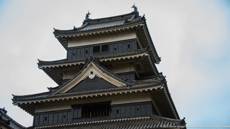 Low-angle view of Matsumoto Castle’s dark upper stories, white gables, barred windows, and tiled eaves.