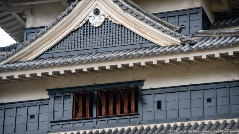 Close view of a white gable, gray roof tiles, crest ornament, black wall panels, and a barred castle window.