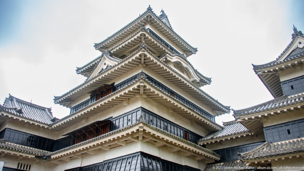Corner view looking up at Matsumoto Castle’s tiered main keep, with broad tiled eaves and dark walls.
