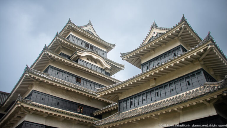 Oblique upward view of Matsumoto Castle’s main keep and adjoining rooflines beneath a cloudy sky.