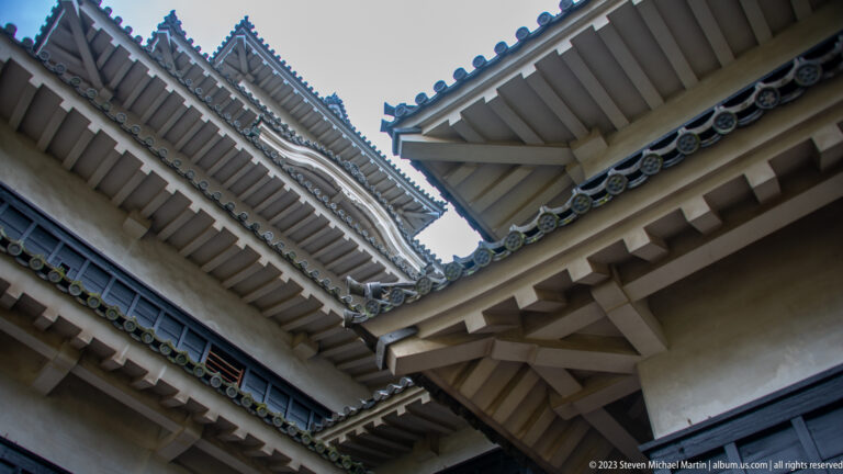 Upward view between intersecting Matsumoto Castle eaves, showing rafters, white plaster, dark walls, and roof tiles.