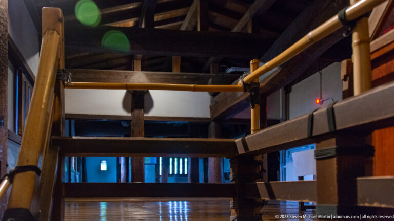 Steep interior stair, dark timber beams, polished floorboards, and visitor railings inside Matsumoto Castle.
