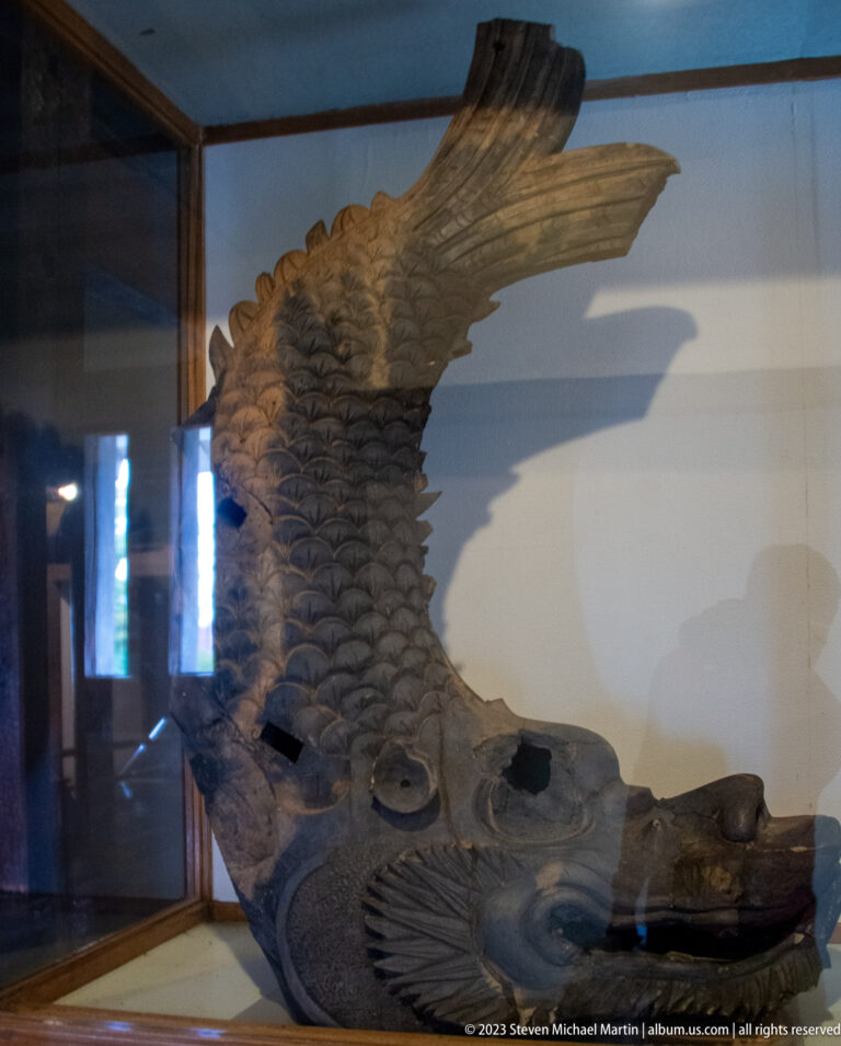 Weathered carved wooden roof ornament with a curved scaled body and extended fin displayed behind glass.