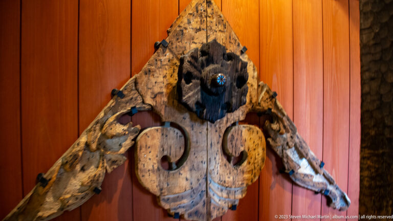 Weathered triangular wooden architectural ornament with a dark central crest and curled lower motifs.