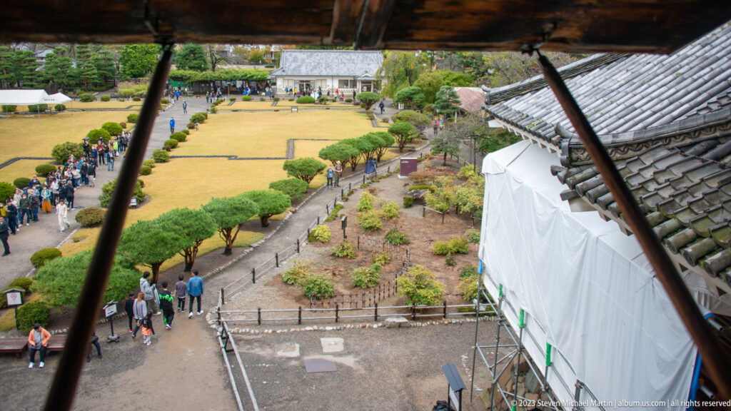Elevated view from Matsumoto Castle over garden paths, clipped pines, visitors, and nearby tiled roofs.