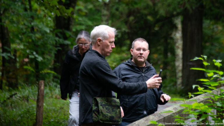 Three adults on a forest path, two men looking at a smartphone in front.
