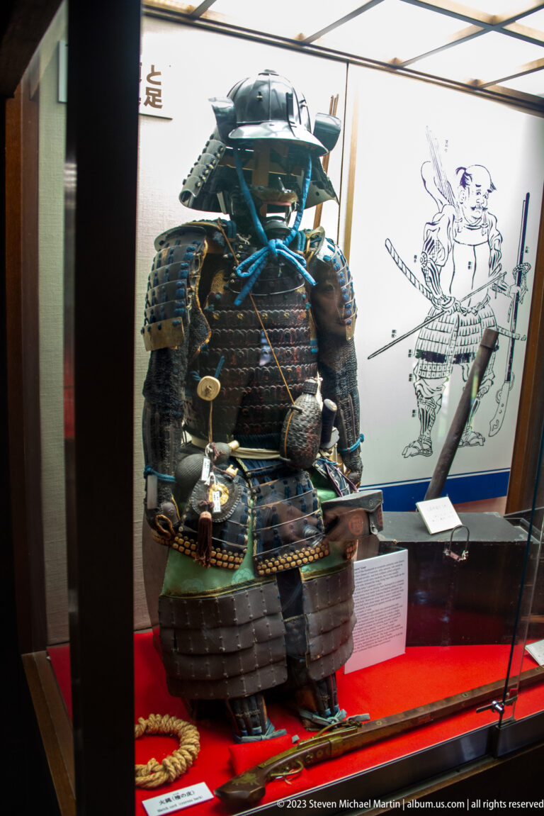 Dark Japanese samurai armor with helmet, face guard, layered cuirass, cords, and weapons in a display case.