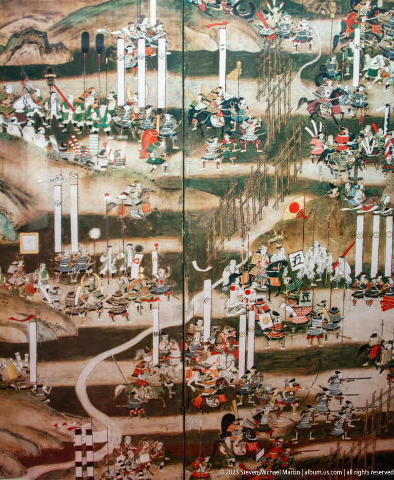 Large Japanese folding-screen painting crowded with warriors, banners, horses, weapons, and encampment structures.