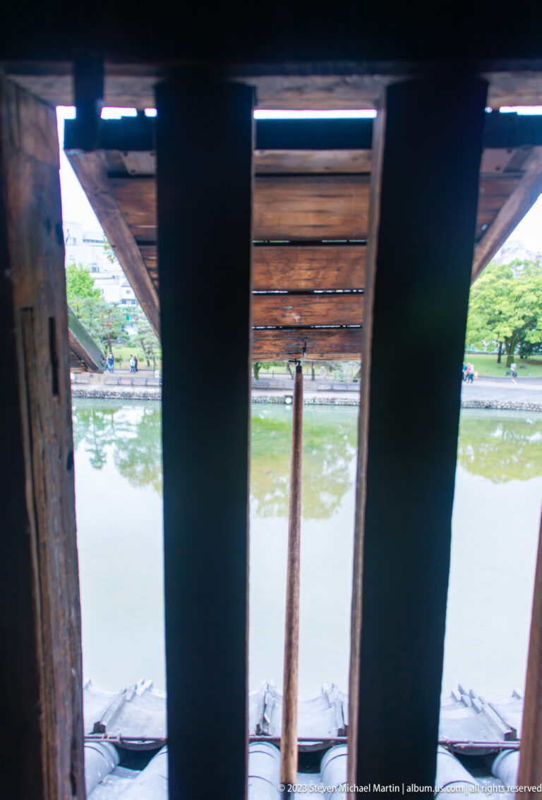 View through heavy timber bars inside Matsumoto Castle toward the moat, trees, and opposite bank.
