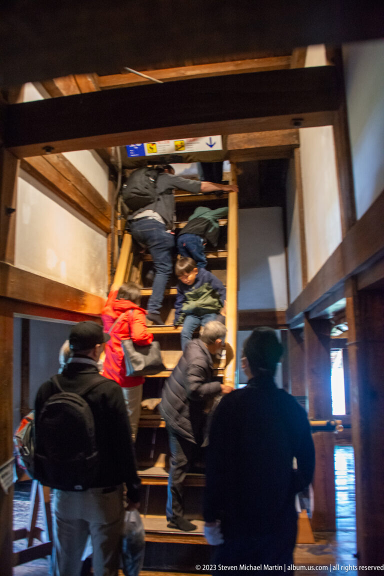 Visitors climbing a steep wooden stair beneath heavy beams inside Matsumoto Castle.