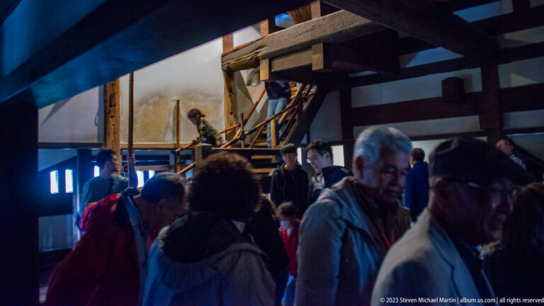 Crowd moving among heavy timber posts, beams, stairs, and ladders inside the dim Matsumoto Castle keep.