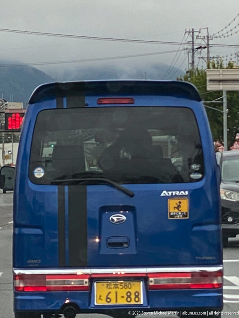 Rear view of a blue passenger van in traffic, with utility lines and mountain slopes in the distance.