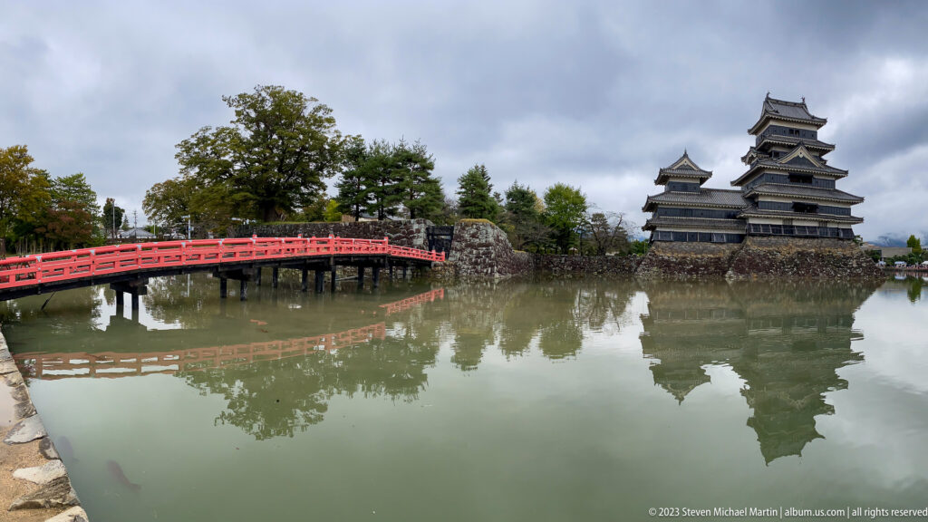Matsumoto Castle across its moat beside a vermilion bridge, with both reflected in calm water.