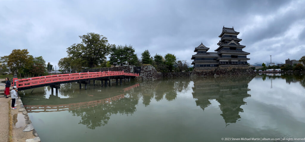 Wide view of Matsumoto Castle, a curving vermilion bridge, trees, and reflections across the moat.