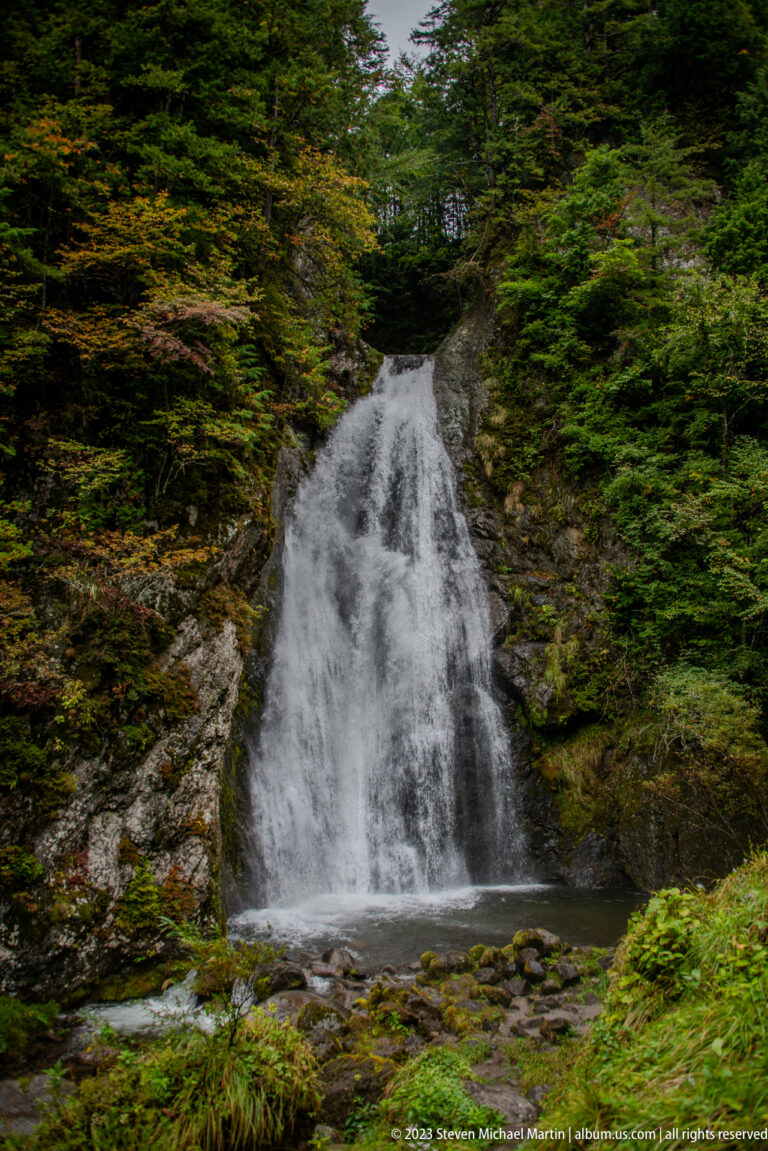 Broad white waterfall descending over dark rock amid dense green forest.