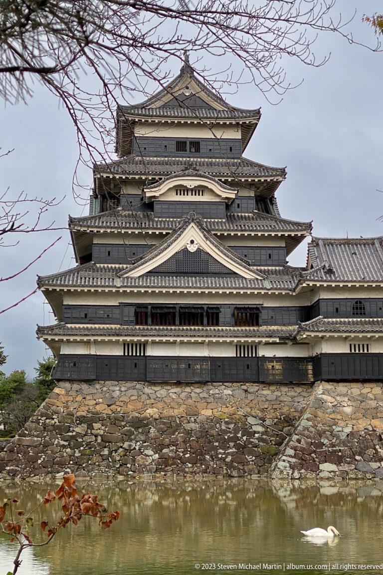 Broad side view of Matsumoto Castle above a stone base and moat, framed by bare branches.