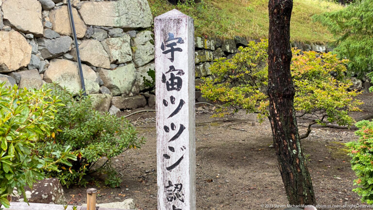 Japanese-inscribed stone marker for Uzumi Bridge beside a masonry wall, shrubs, and a pine tree.
