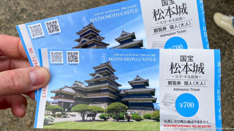 Hand holding two blue Matsumoto Castle admission tickets printed with the castle image, National Treasure label, and 700-yen price.