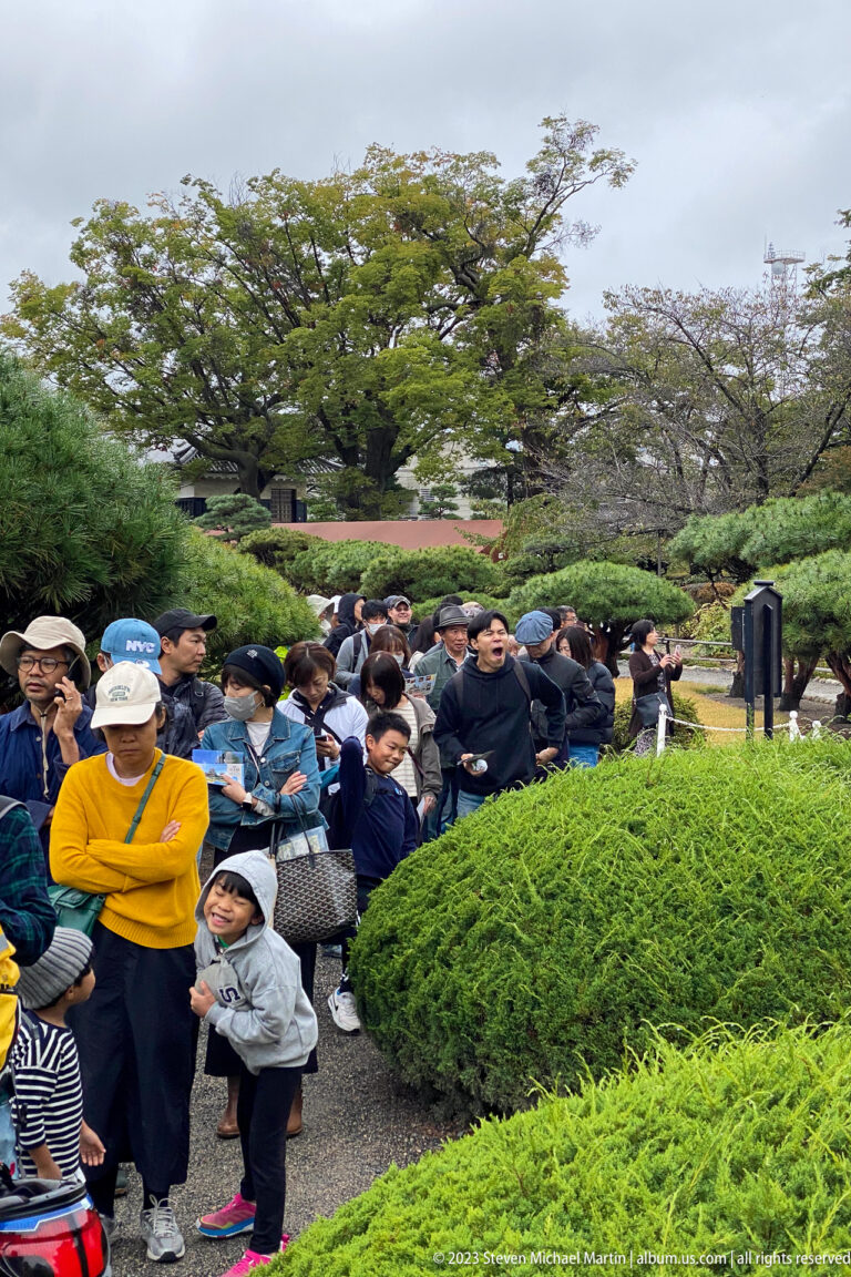 Adults and children queue along a path between clipped green shrubs and trees in the Matsumoto Castle grounds.