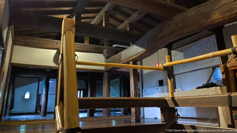 Heavy wooden posts, crossbeams, roof members, and a visitor railing inside a dim Matsumoto Castle landing.