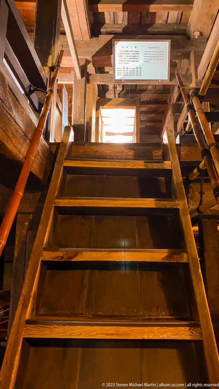 Steep ladder-like wooden staircase rising through heavy beams toward a bright upper opening inside Matsumoto Castle.